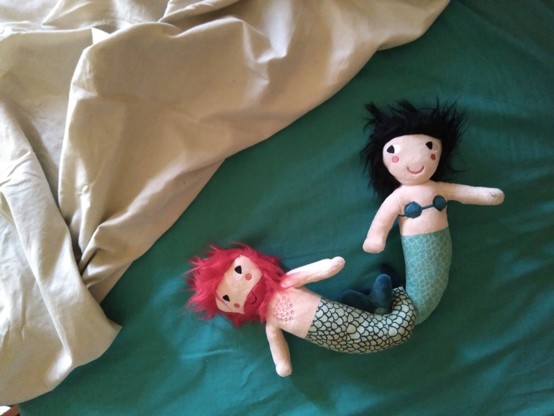 Un sereo e unha serea de peluche nunha cama con sabas brancas e azuis. Teñen as colas enlazadas.